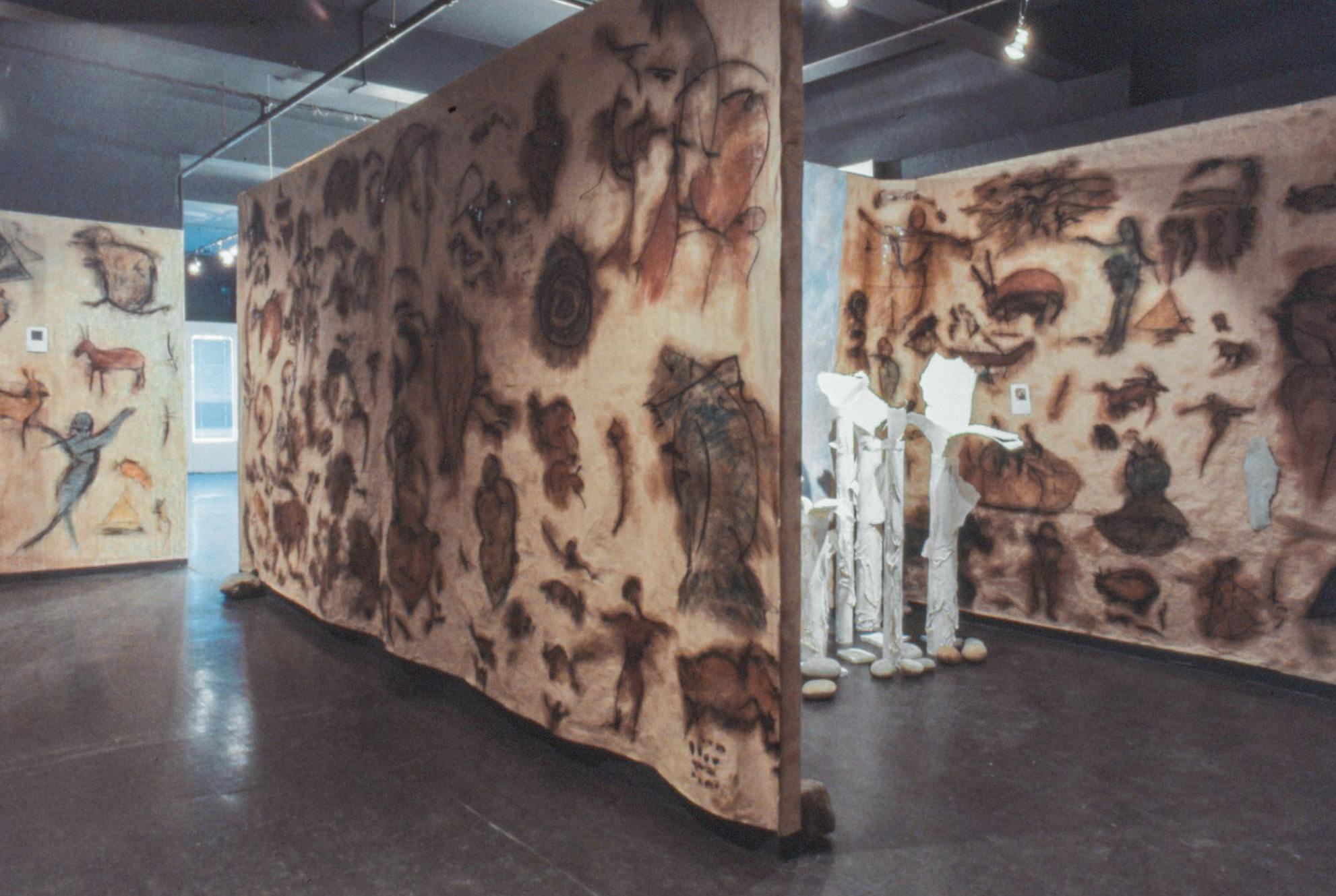 A gallery space with walls covered in pale brown paper that has chalky drawings of animals and people. One area is sectioned off by walls and tall white sculptures are partially visible.
