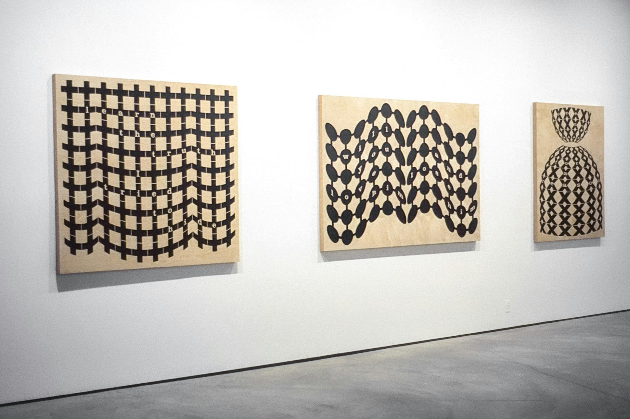 3 artworks on wood panels mounted on a white wall. They show black geometric patterns on natural wood with text. The work on the left has a grid pattern that reads "learn the truth I tried to hide." 