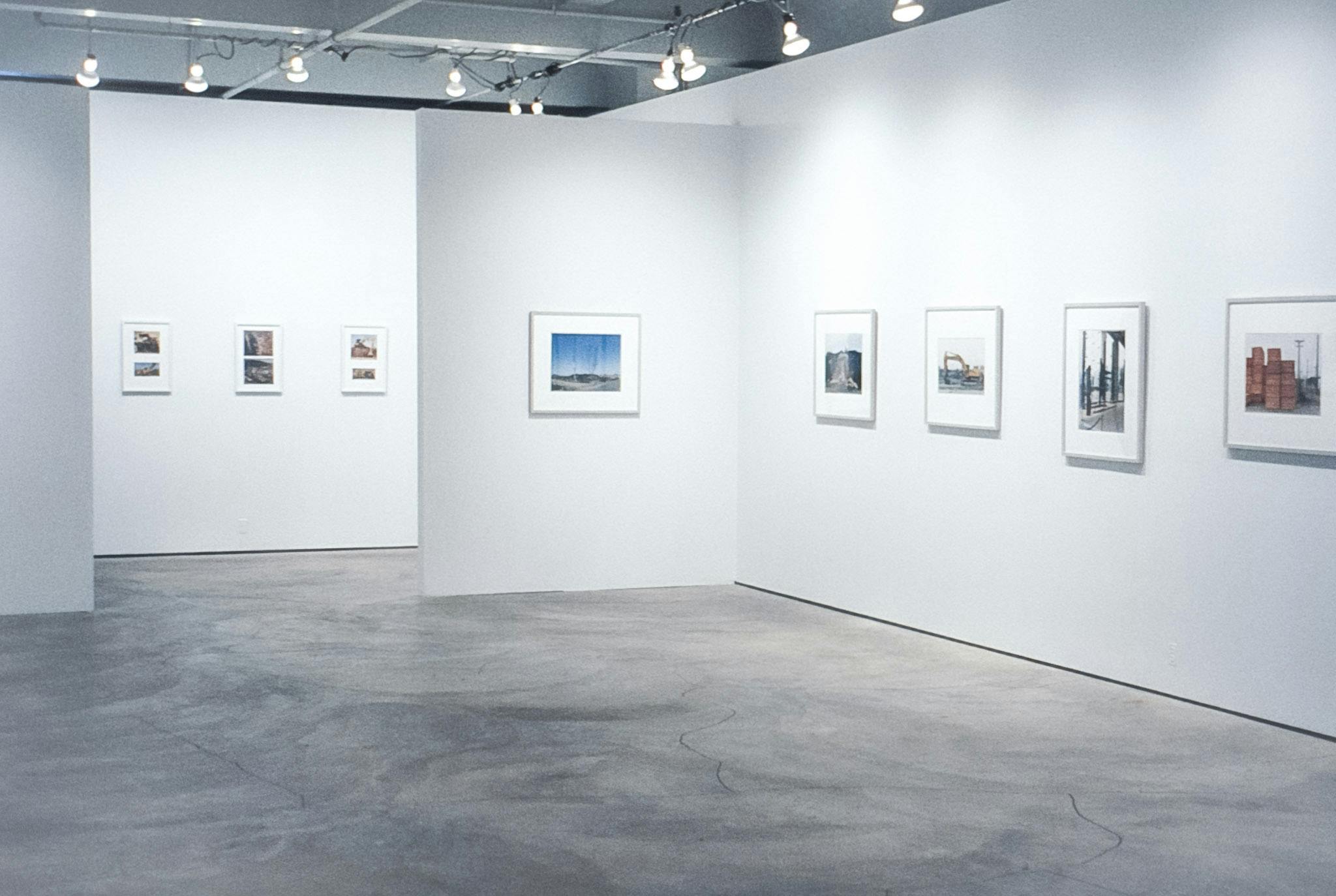 The walls of a gallery space and a hallway. On all walls, there are framed photo works. The frames in the hallway contain 2 photos, and the frames in the interior walls have 1 larger photo in them. 
