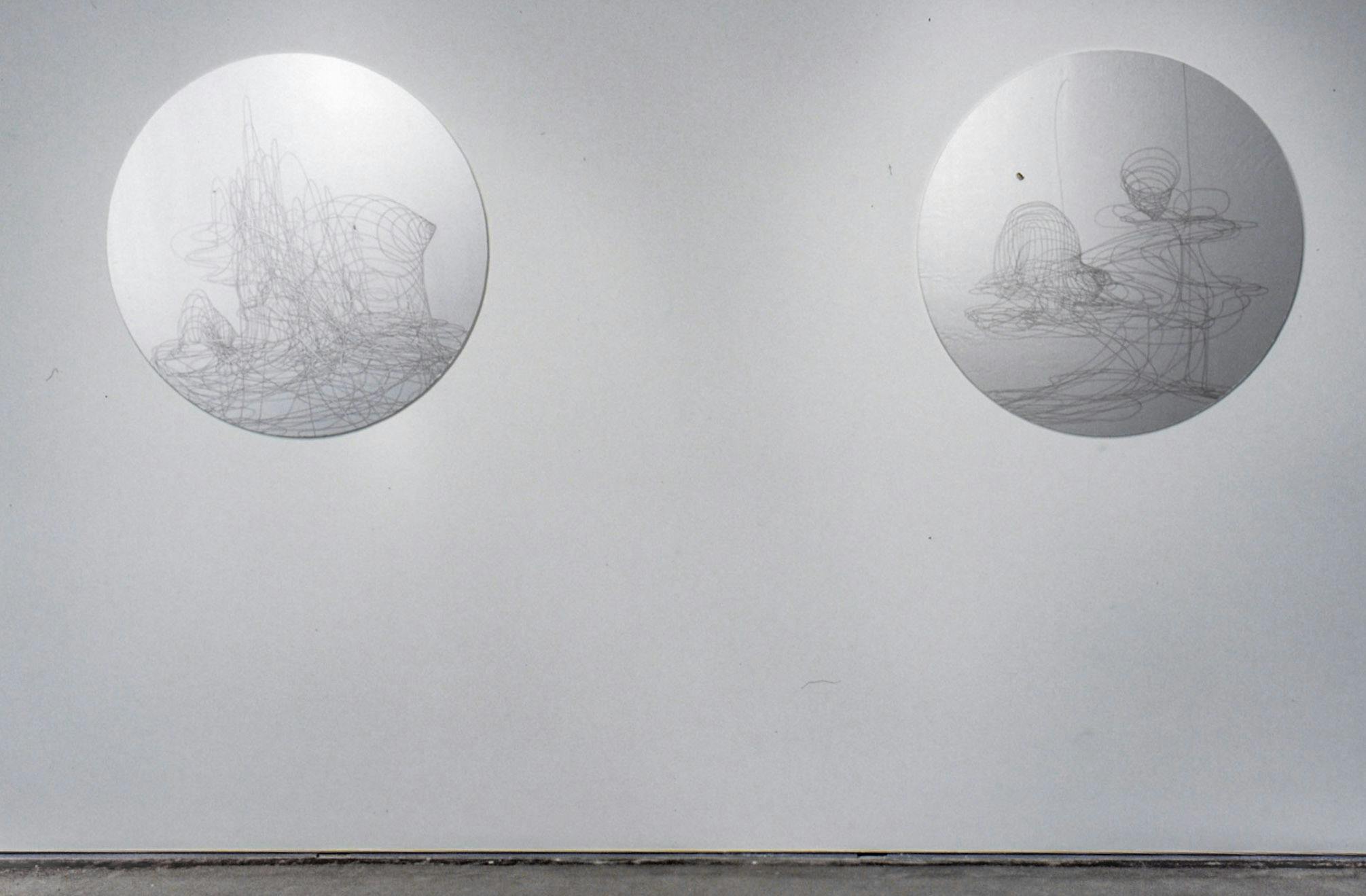 Two circular shaped sculptures are mounted on the gallery wall. They both have a silver metallic surface, on which some abstract line drawings are made. 