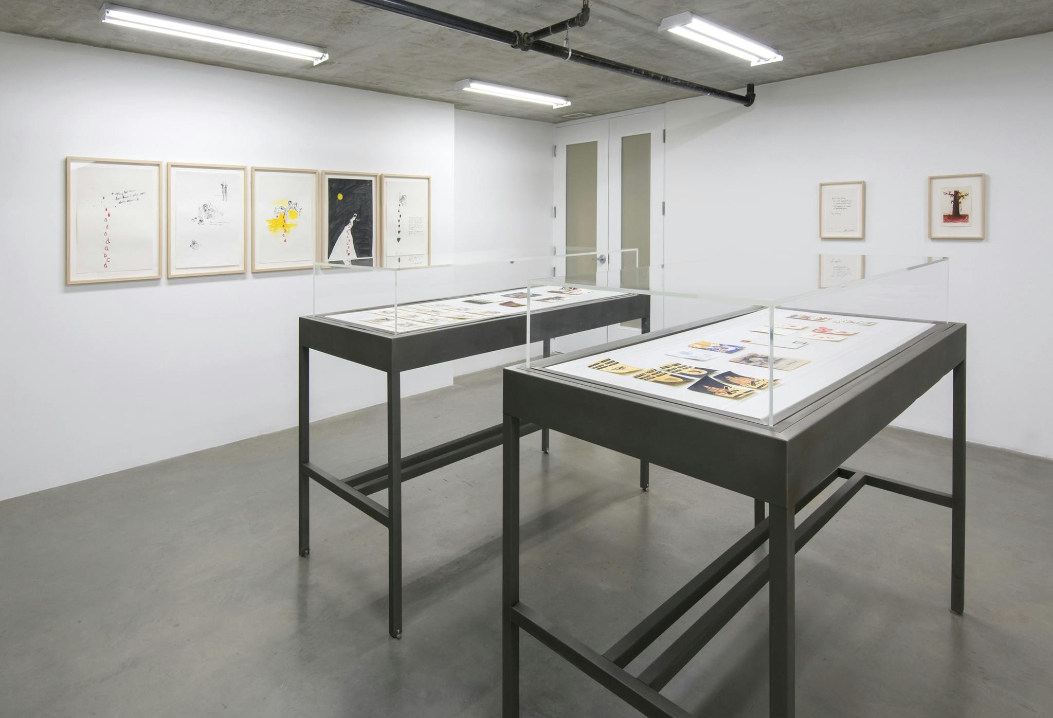 Installation image of eight framed works on paper hanging on a wall. Two vitrines sit at the center of the room within which various small works on paper are displayed. 