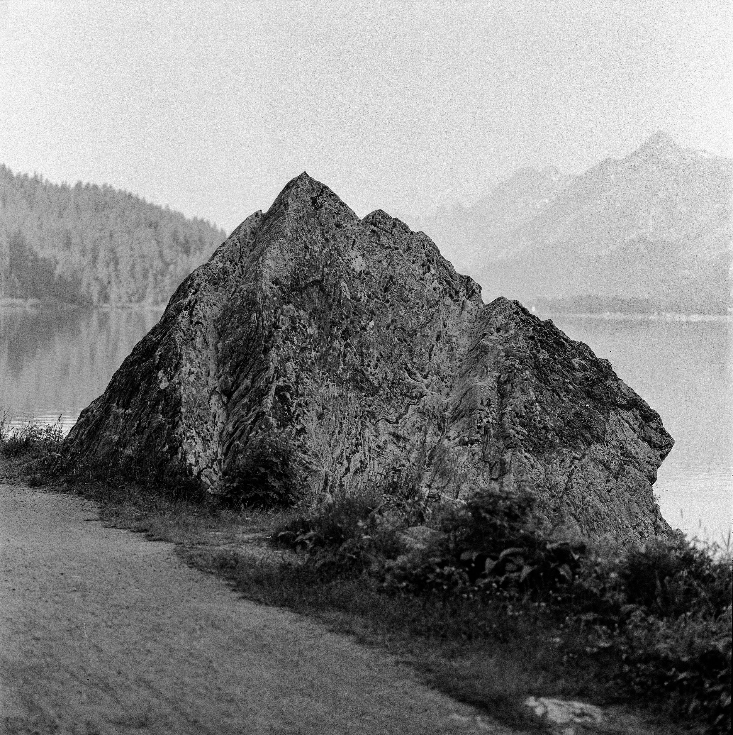 A black and white photograph depicting a large triangle-shaped stone on the shore of a lake, with mountains faintly visible in the background.