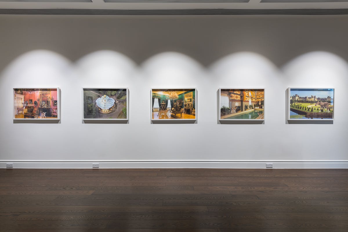 Five framed images hang on the wall of a gallery space. The images display a variety of lavish indoor and outdoor spaces, with irregular animals, like a tiger and a moose, existing within these spaces.