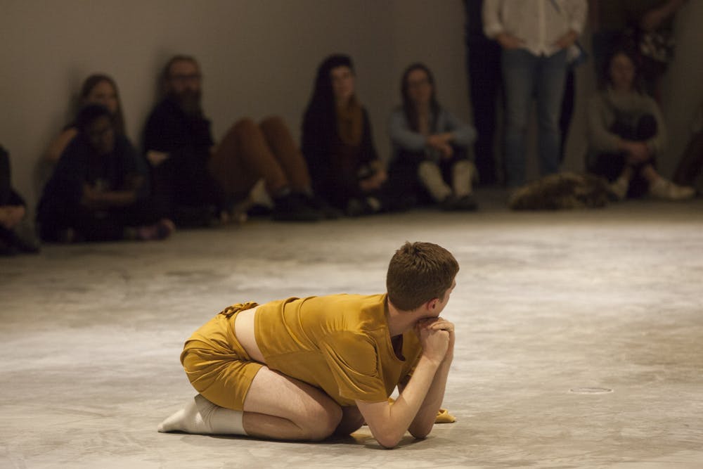 A performer dressed in yellow sits on the floor of a gallery space with audience members standing around the perimeter. They are curled up on their knees, propping their head up with their hands.