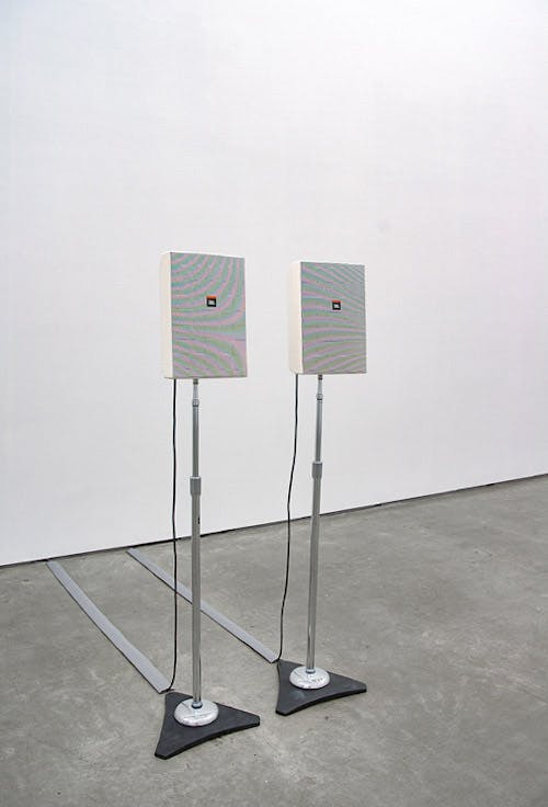 An installation view of a work titled Stereo by Ceal Floyer. Two identical speakers on tall stands are placed in the middle of a gallery space. 