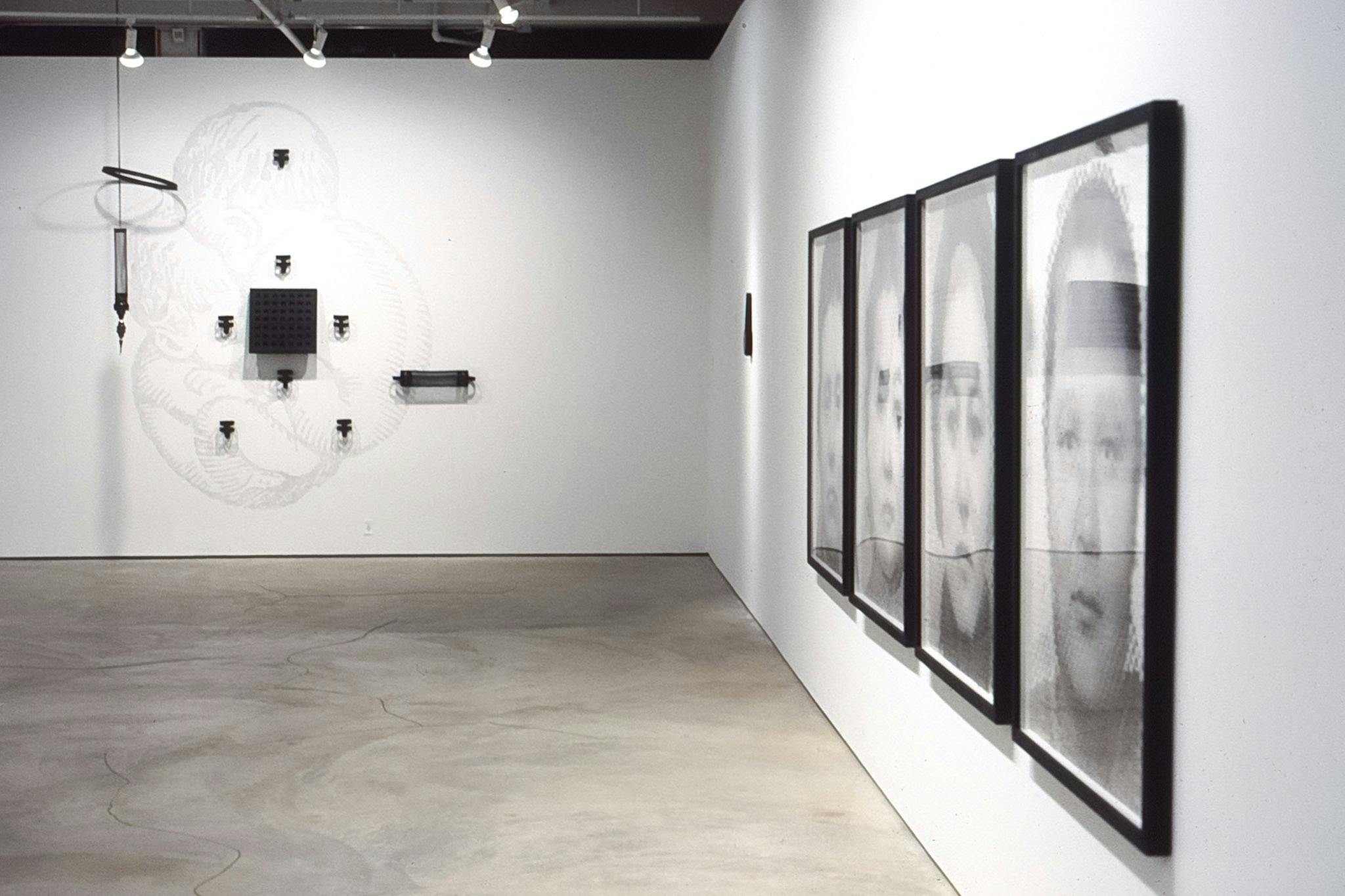 An installation view of the gallery shows four black and white portrait photographs on the right-side wall. On another wall, a group of black sculpture works are mounted. 