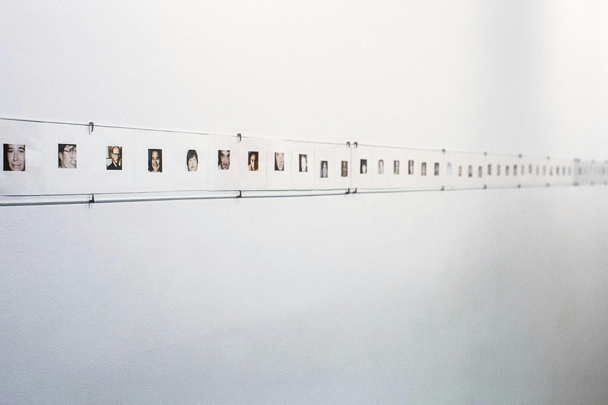 A photo of a photo collage installed in a gallery wall. The photographed faces of different people align horizontally in a long stretched white sheet of paper mounted on a white gallery wall.