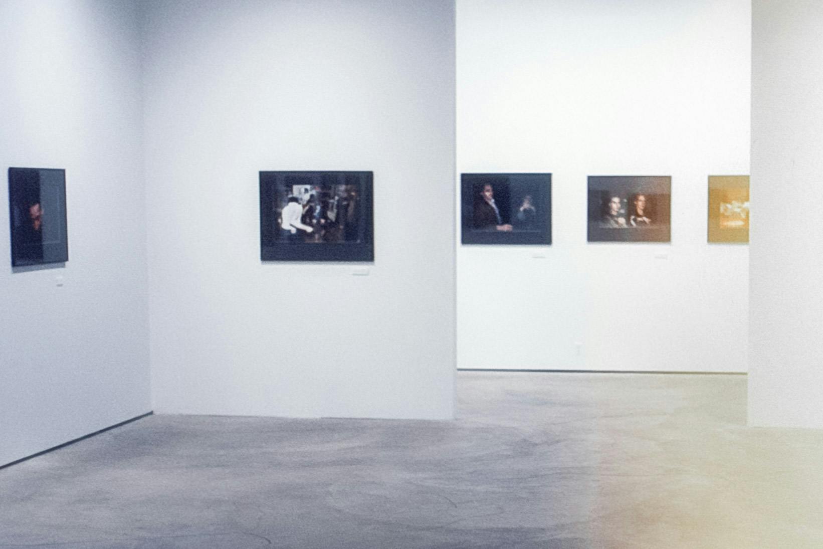 The corner of a gallery space with a hallway visible in the background. On each wall, there are photographic works in black frames. The photos show people dancing or sitting in dark rooms. 