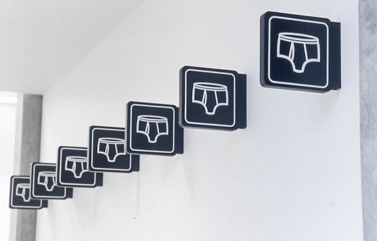 On the upper part of the galley wall, seven identical signs are attached in a row. They are black square-shaped signs. A drawing of white brief underwear is printed on each of those black signs.  