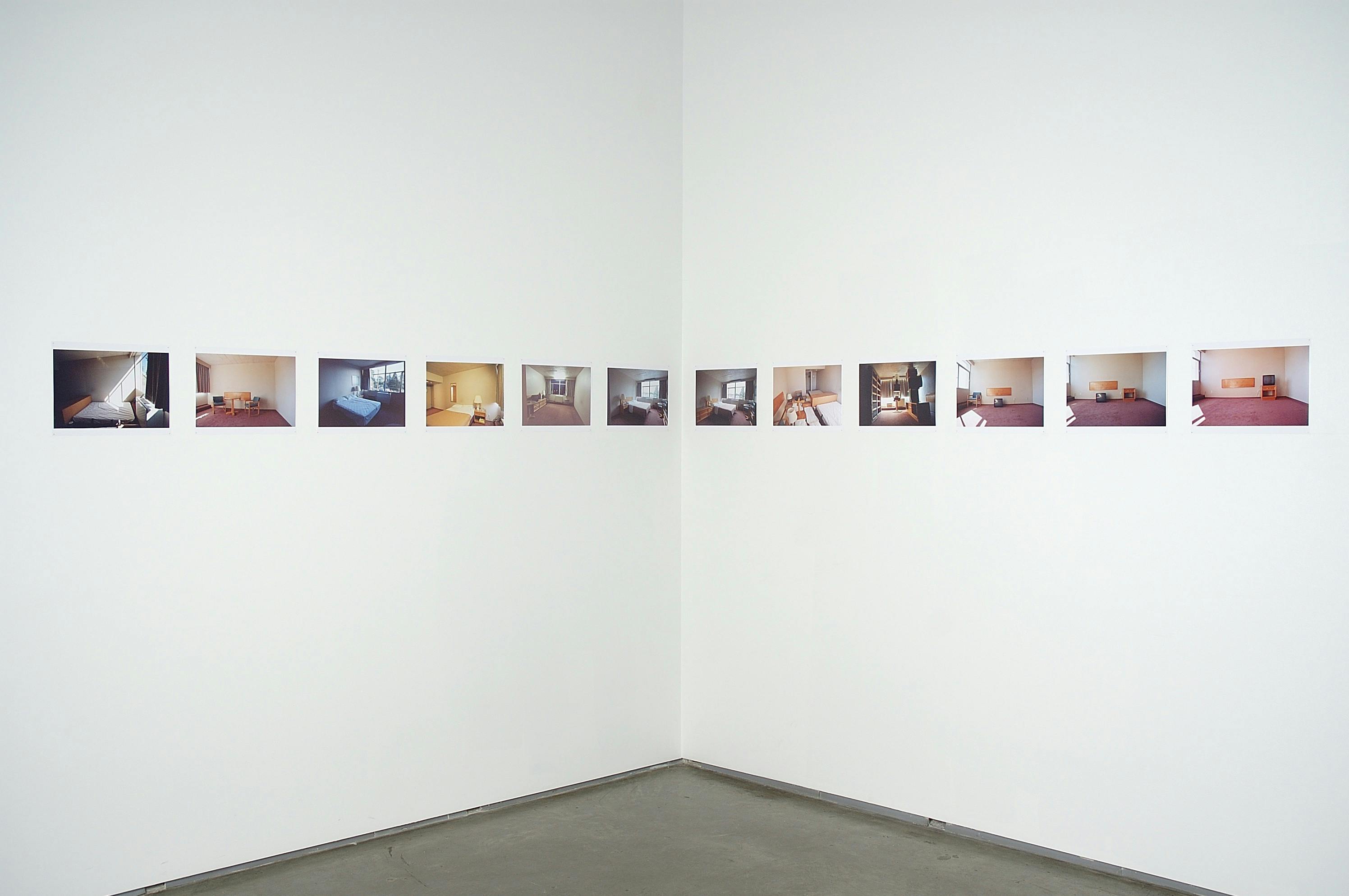 Twelve small, unframed photographs are hung in a line across the corner of two gallery walls. The photographs are of various interior, domestic-like spaces.