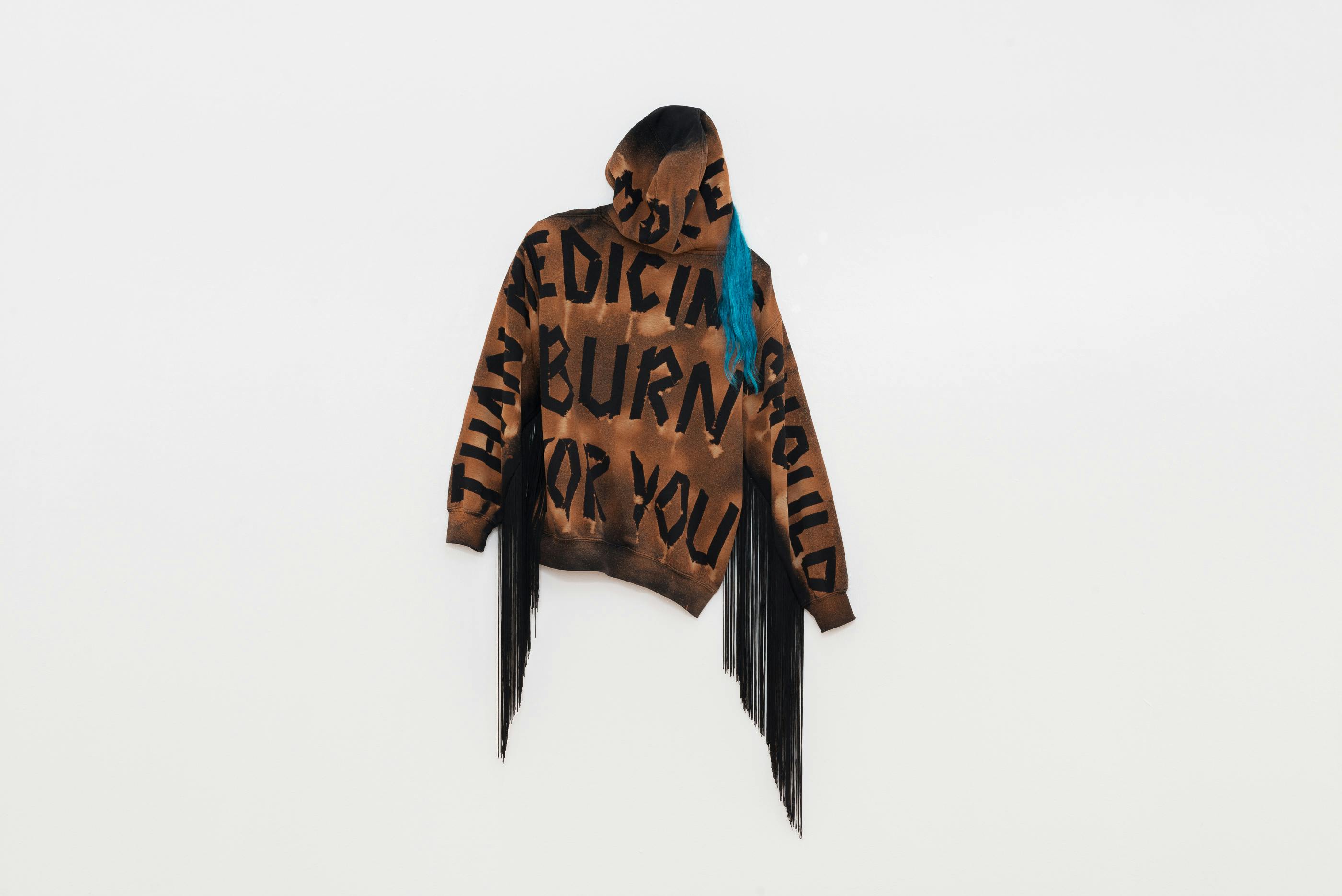 A brown hoodie with bold, black text hangs on a gallery wall so that the back is visible. Blue hair emerges from the hood and black fringe hangs down from each sleeve.