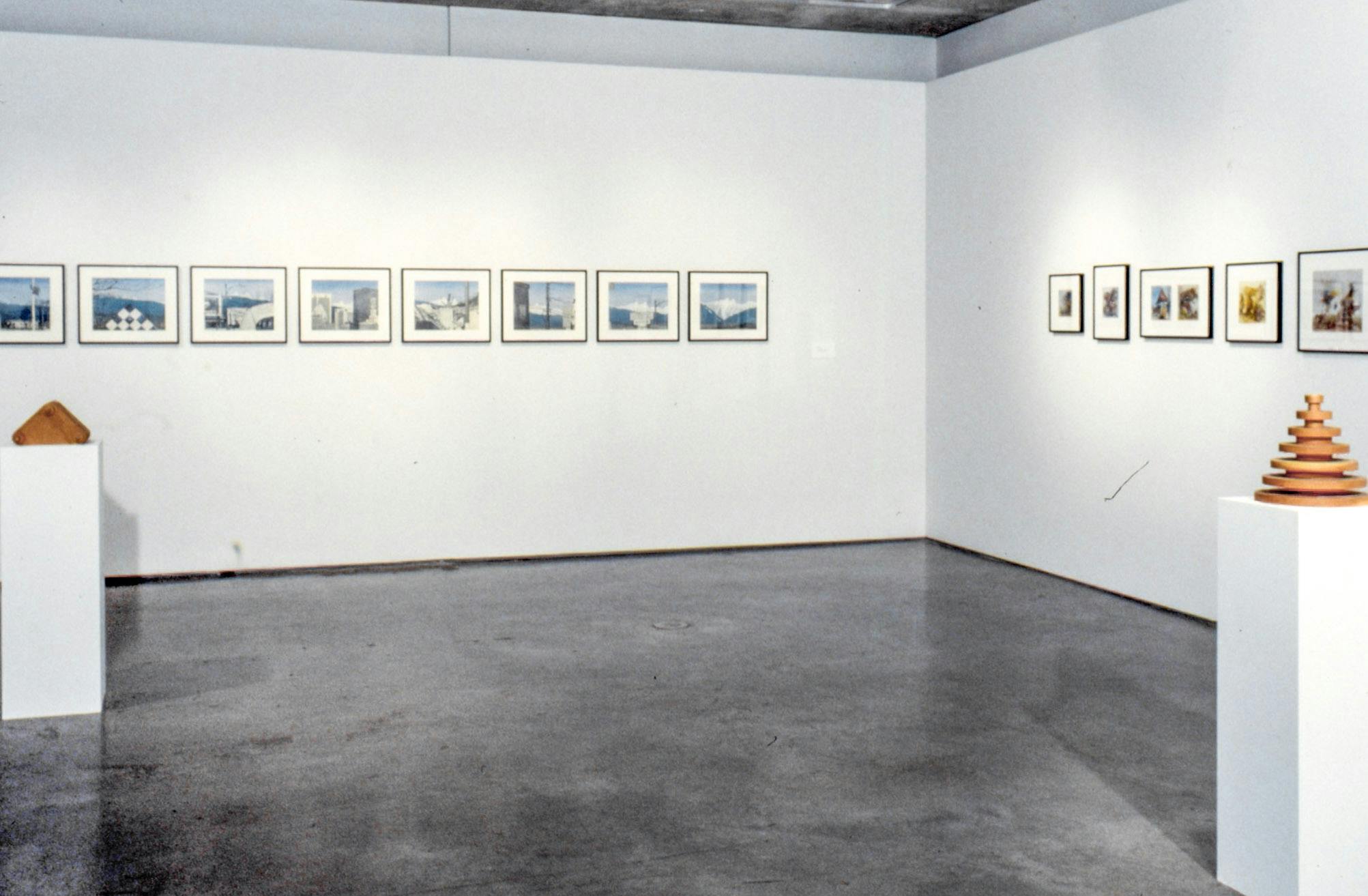In a gallery space, a set of blue and white landscape drawings and another set of small coloured drawings are mounted on the wall. Two wood sculptures are placed on pedestals.