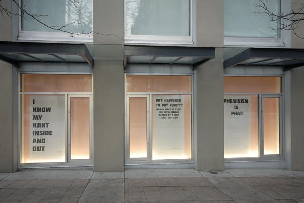 Three large fliers are installed in the CAG’s window spaces. They are all text-based works showing slogans such as “Pheminism is Phat.” All the walls behind those windows are pink. 