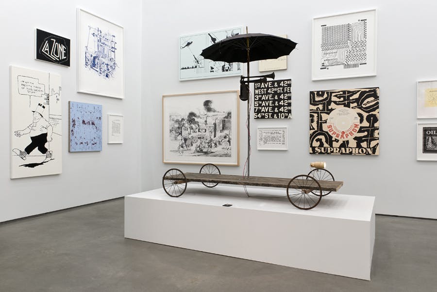 Various artworks are installed in the corner of a gallery. Multiple two-dimensional works of different sizes cover the walls behind a large-scale sculpture installation resembling a cart.