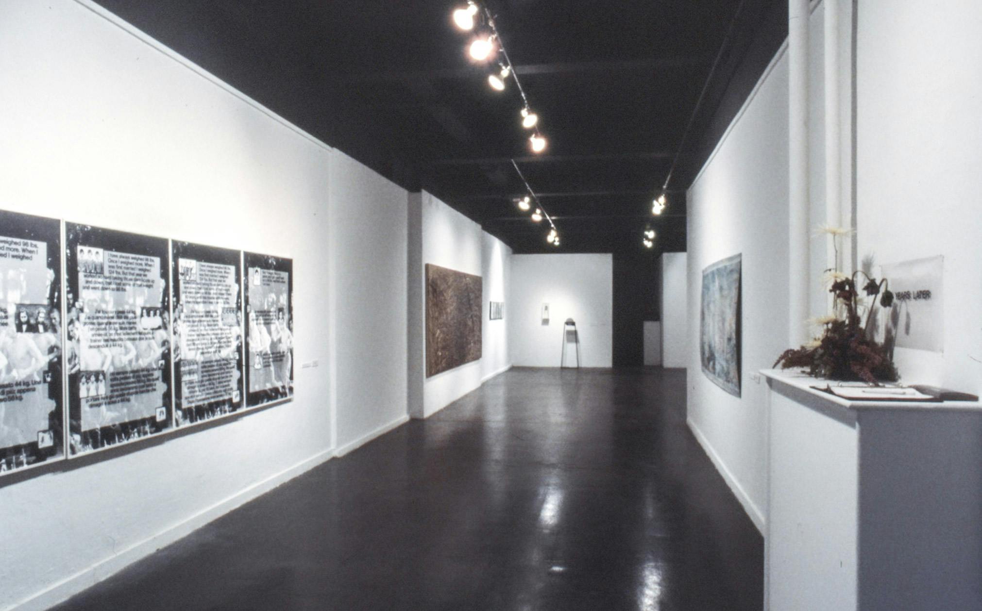 Several artworks visible in the hallway of a gallery. The works include large abstract paintings on canvas and paper, large framed photos of a group of people with text overlay, and small sculptures. 