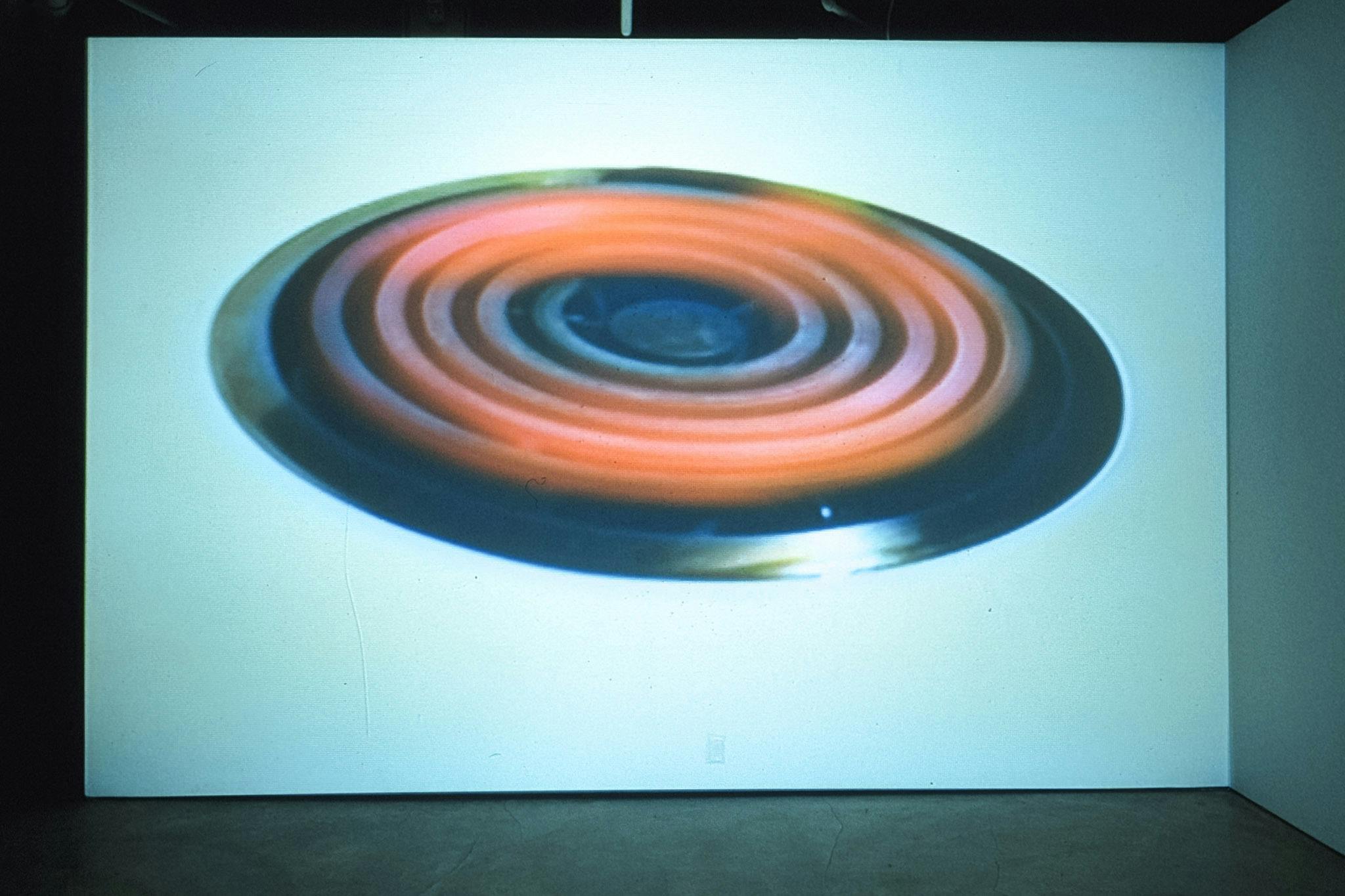 The large oval shape is projected on the white gallery wall. It is a cut-out image of an electric coil stove for cooking. The stove’s orange colour suggests that it is generating heat.