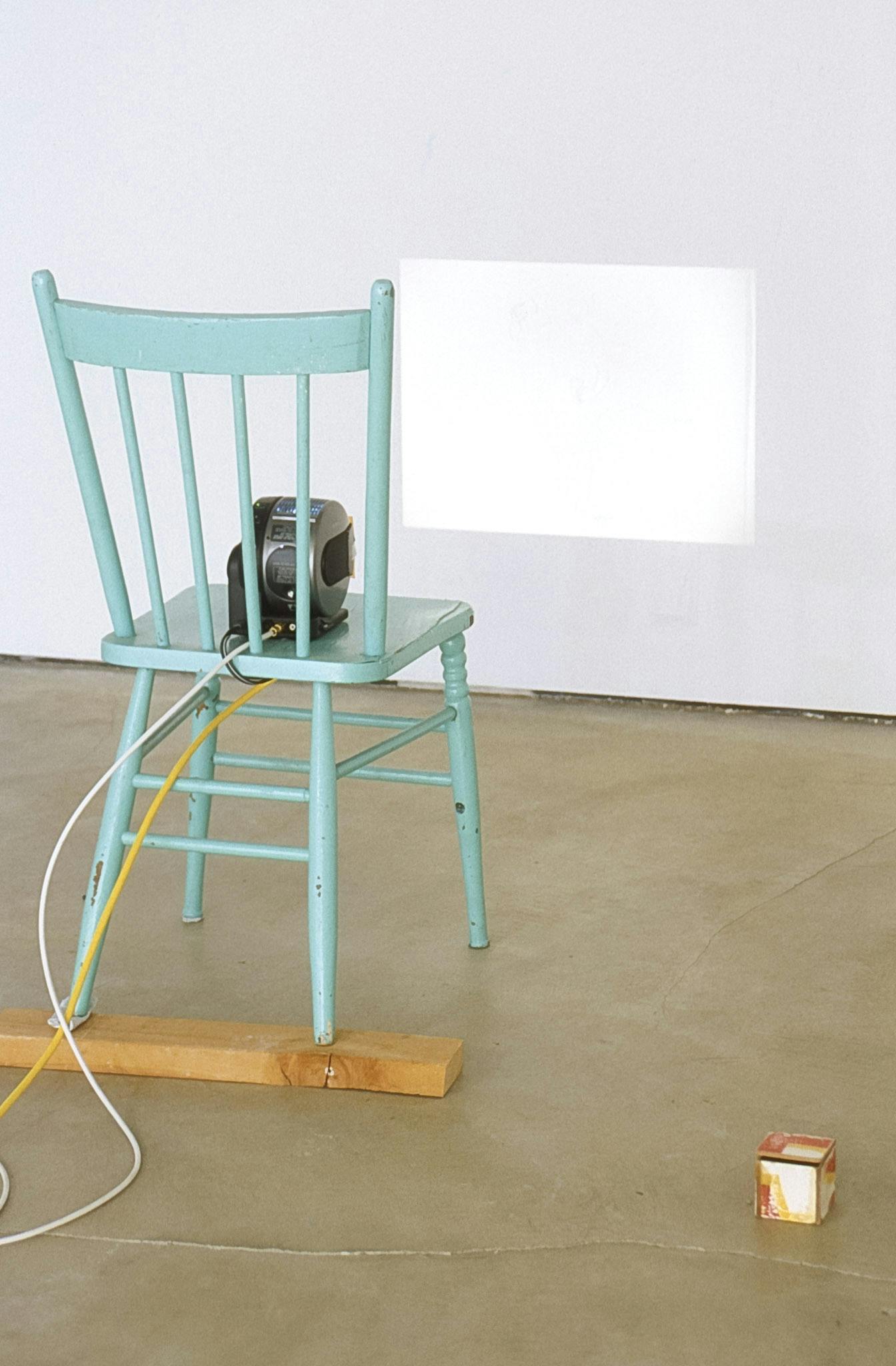 A small projector is placed on the light blue painted wooden chair in the gallery. The projector, connected to white and yellow codes, projects a blank white rectangle on the white gallery wall.