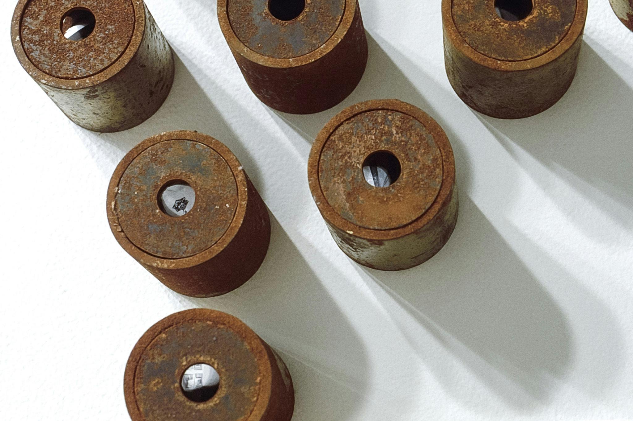 Six short cylinders are visible in this image. They are made of rusted metal. Viewers can peek at the black and white image at the bottom of each cylinder through the hole on top of each cylinder.  