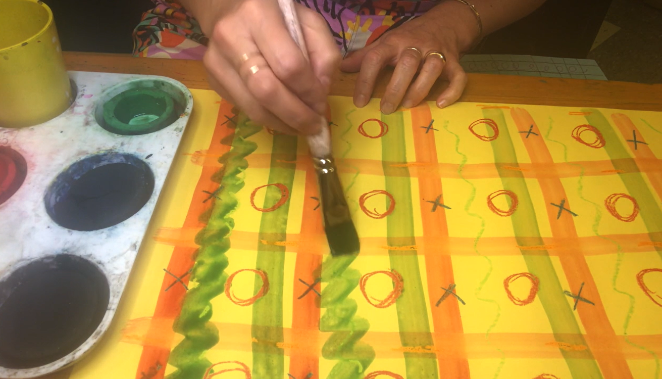 A still of a hand painting with a paintbrush. On a large sheet of yellow paper, a red grid, red circles, and green lines are drawn. 