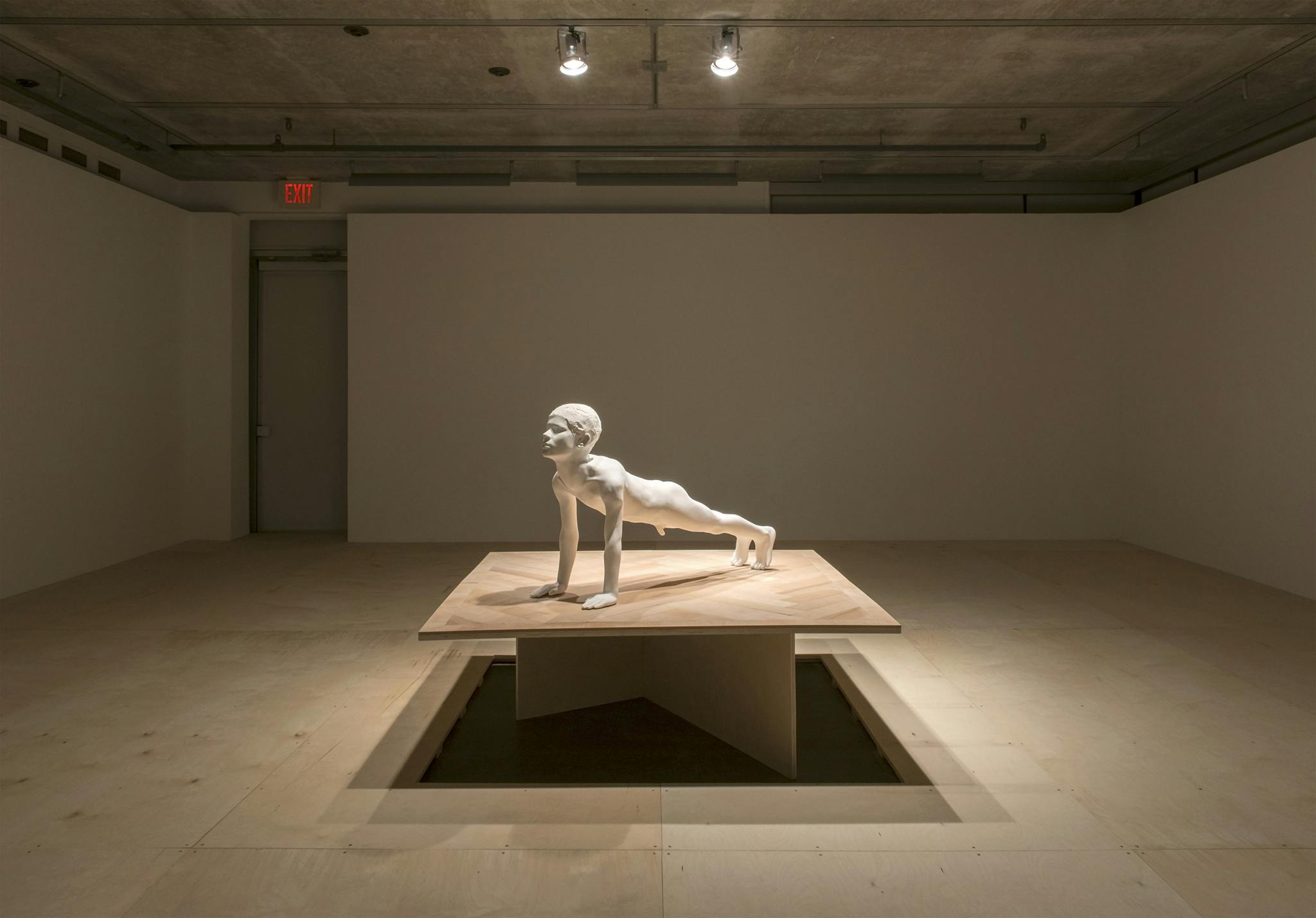 A white sculpture of a male nude in a plank position installed in the middle of a gallery. The sculpture is at human scale and installed atop a wooden plinth. 