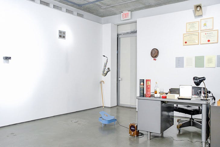 An installation image of a work by Daniel Olson. A steel-made grey working desk is placed directly on the floor. The desk is covered with various objects, including a typewriter, a desk lamp, and a bottle of rum. 