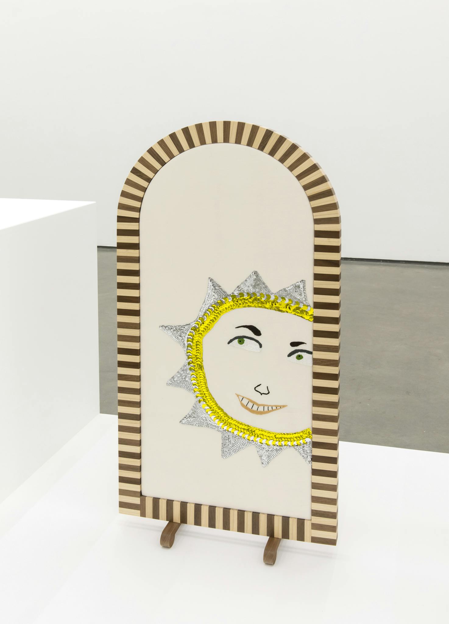 A fire screen with a brown and beige striped wooden frame sits on top of a plinth. Within the screen, a thread and sequined embroidery sun with a grinning face sits off center. 