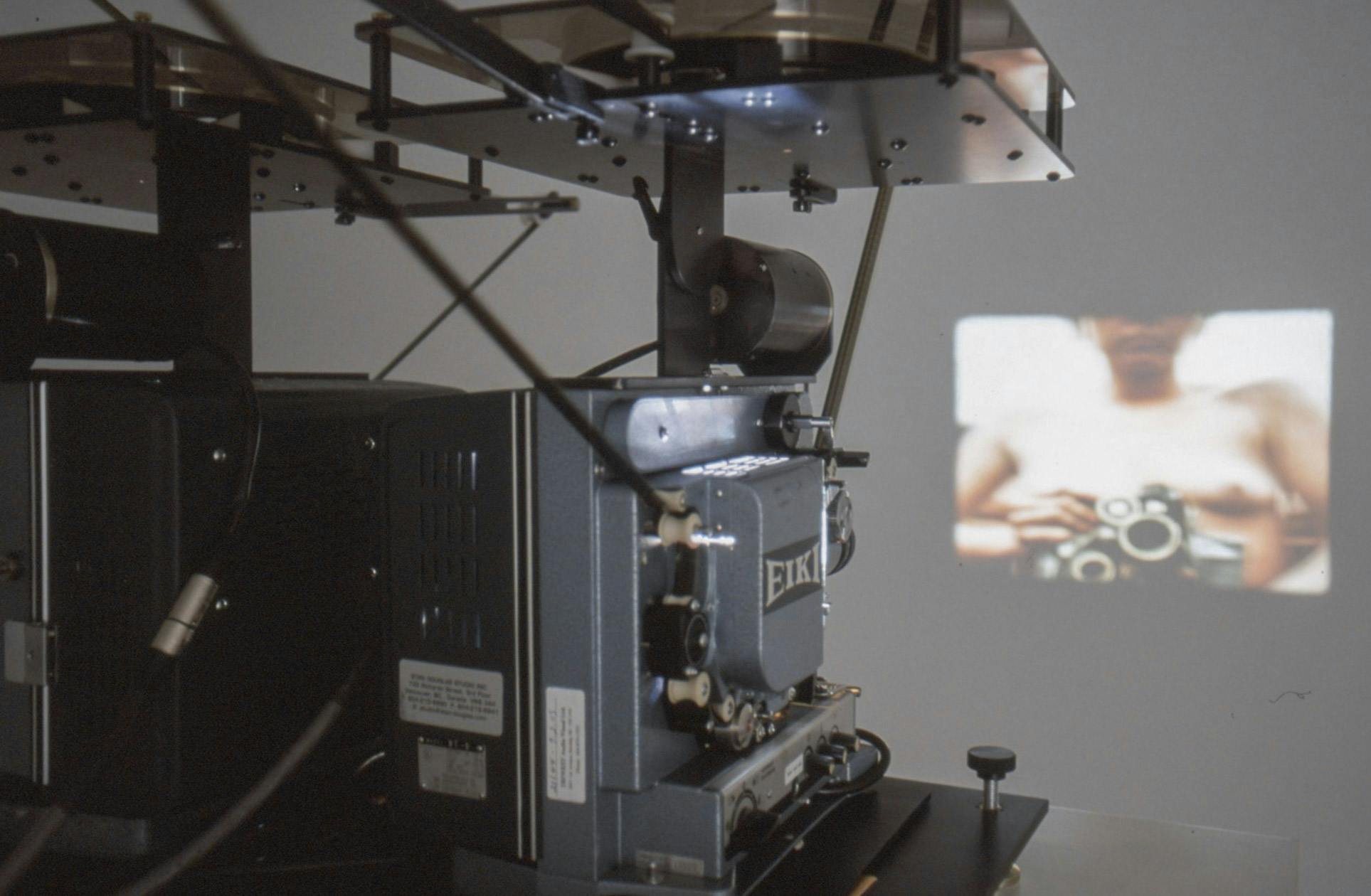 A closeup showing a film projector playing a video on a wall. The video shows a naked person sitting with a camera in their hands, filming.