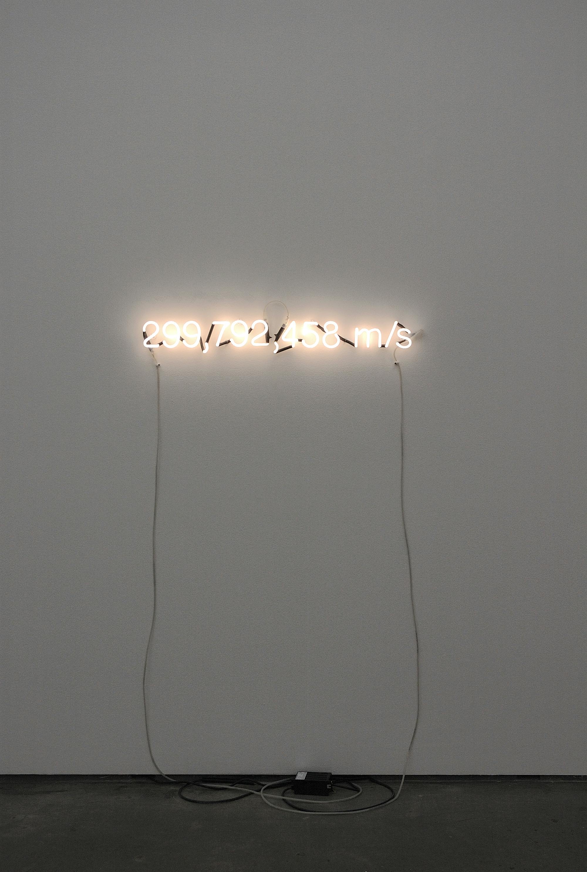 A white neon sign is attached to a white wall. It is attached some feet above the floor. The sign reads “299,792,458 m/s.” White tubes are hanging from both ends of the sign.