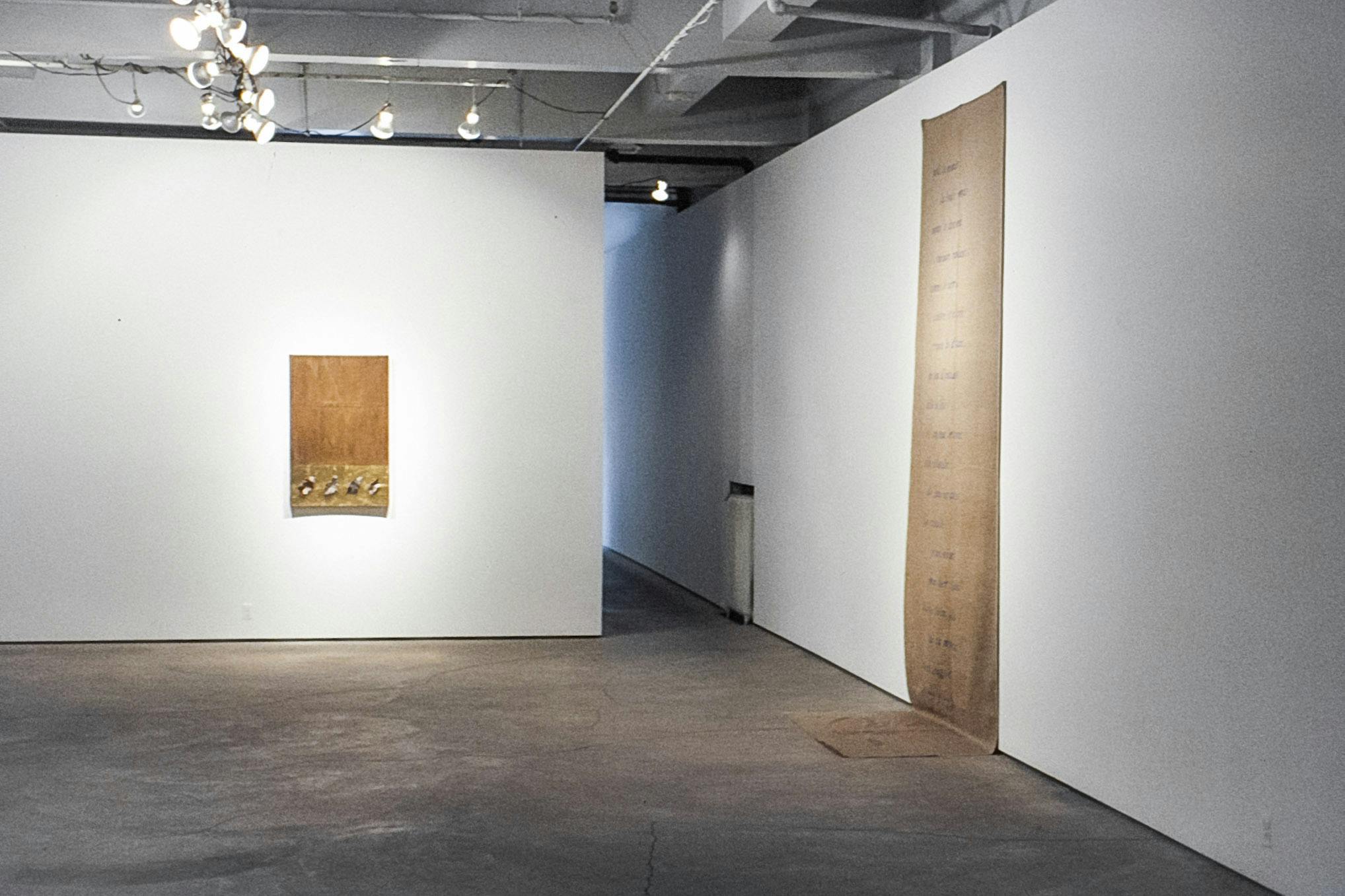 A gallery with one artwork on each of the visible walls. One work is a long piece of fabric that touches the floor. One is a wood panel with gold foil and small bits of material along its bottom edge.