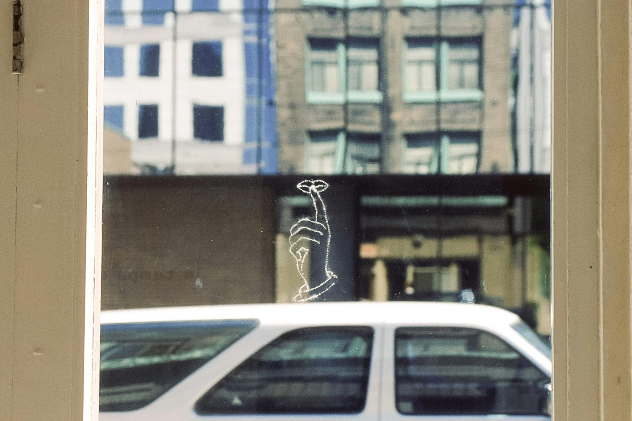 A photograph is attached to the beige-painted door. The picture shows a building’s glass facade behind a passing car. A white line drawing of a finger over lips flotes above the car’s rooftop. 
