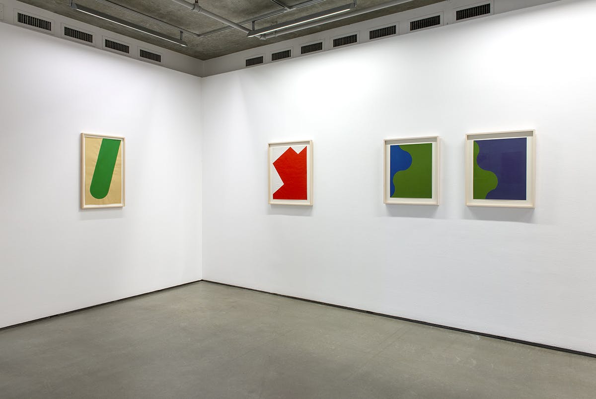 Four framed paintings on paper by Leon Polk Smith sit on either side of a corner in a gallery space. The paintings depict shapes in red, blue, white and green.