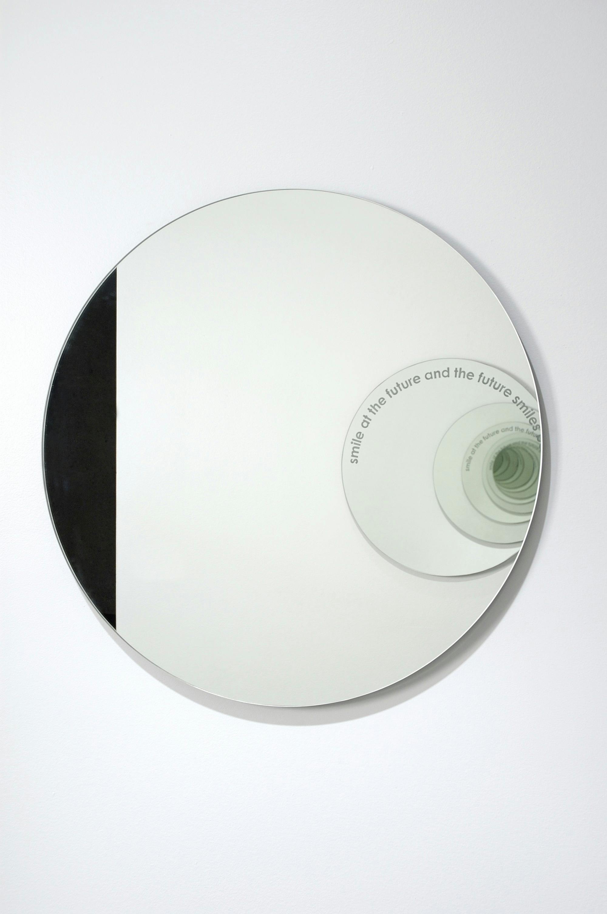 A circular-shaped large mirror is mounted on a gallery wall. It reflects another circular mirror attached to the facing wall. There is a text printed on another mirror.