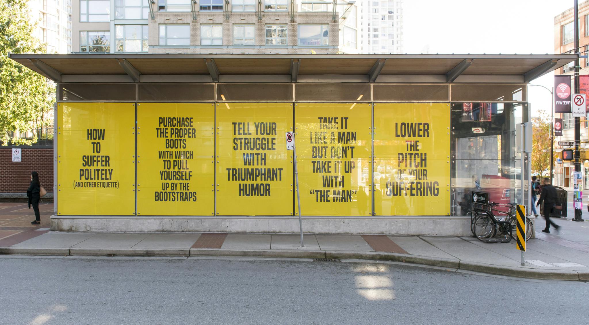The exterior of Yaletown-Roundhouse Station with the work of Kameelah Janan Rasheed installed on the facade. There are large, safety-yellow vinyl prints with large black text on five window panels.