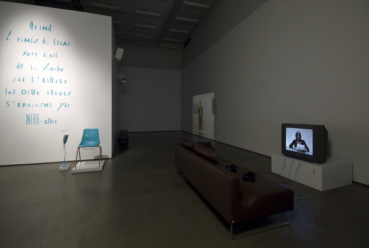 Installation shot of the group show Paying Homage. There is a couch facing a tv, a projection playing directly on the wall, and another wall covered with blue text and a blue chair sitting in front. 