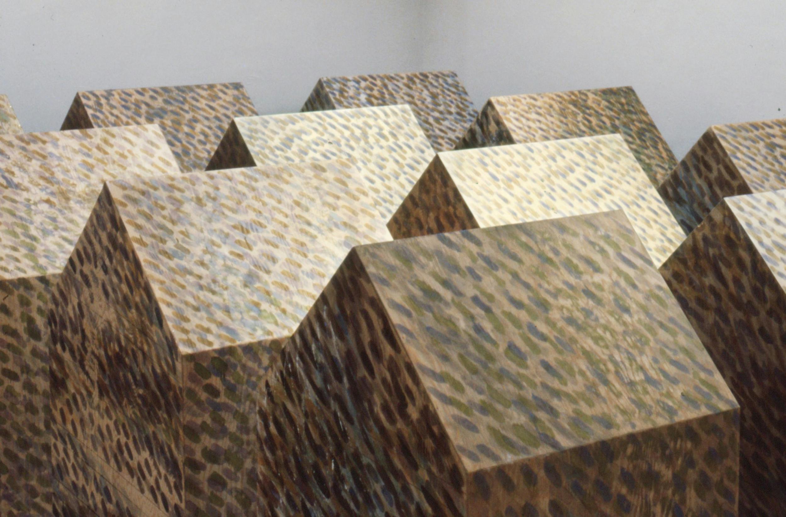 A close up view of wooden sculptures resembling houses are lined up in a gallery. The sculptures are painted with small colourful brush strokes.