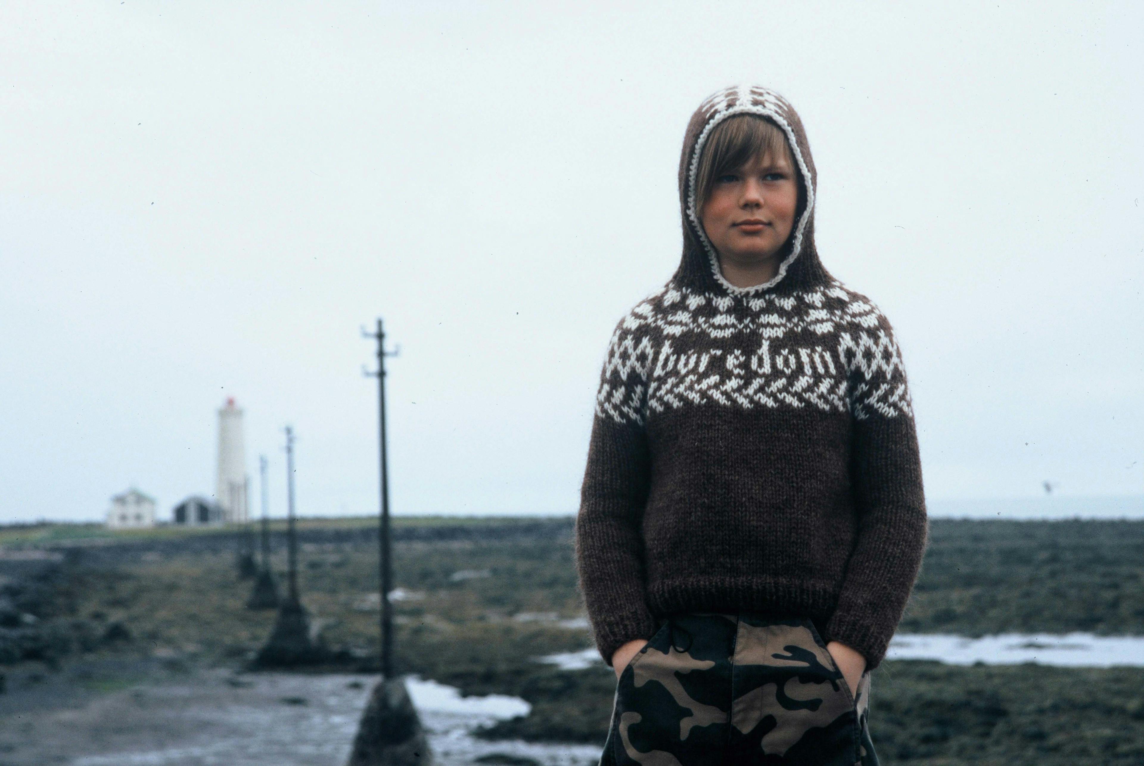 An image of a child wearing a brown sweater facing towards the camera, but looking away, with their hands in their pockets. The blurred background is a stark landscape with buildings in the distance.