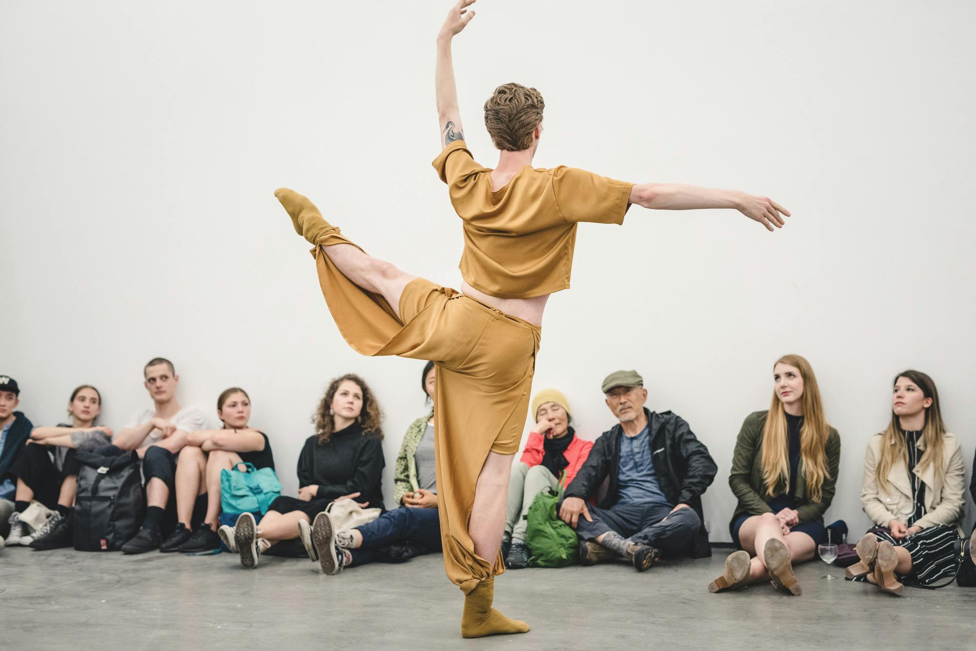 Andrew Bartee wearing a mastered-coloured outfit is posing for the audience in a gallery space. Audience sitting by the wall are looking at this artist’s ballet dancing.