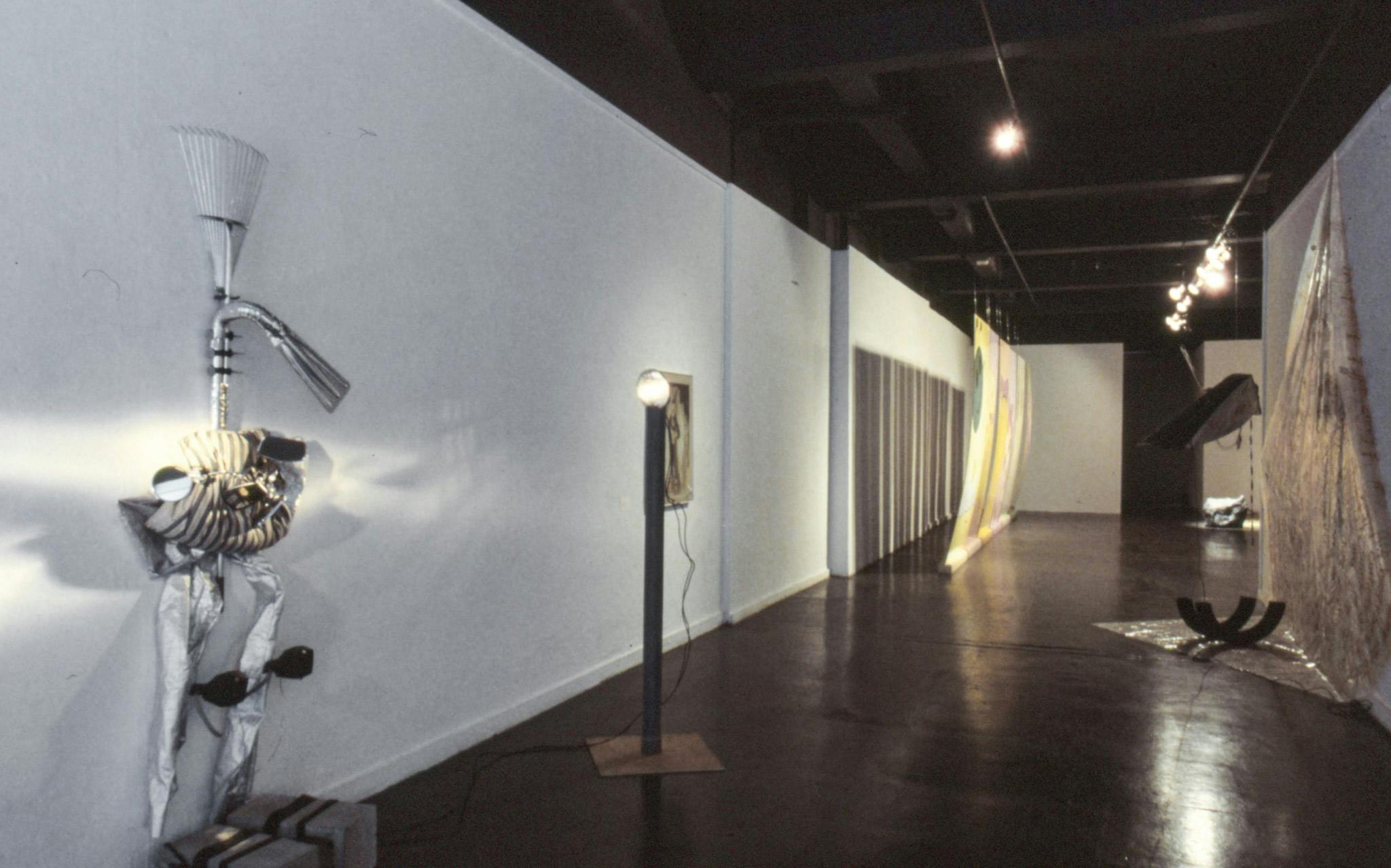 Several artworks in the ball of a gallery. The works include a sculpture with concrete, tin foil, and a metal rake; a sculpture with a lightbulb on a pole; and a painting on the wall. 
