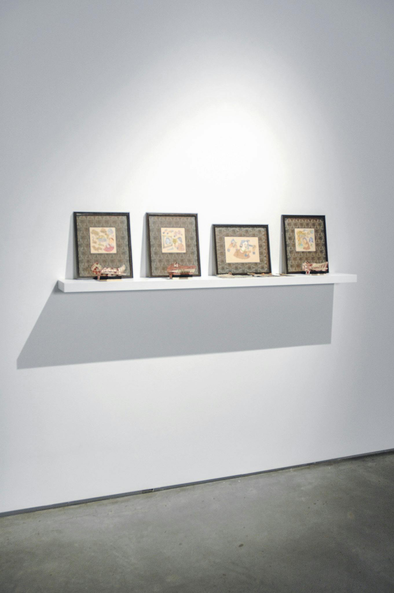 An installation image of artworks in a gallery.  Four coloured illustrations are placed on a small shelf attached to a wall. Small red sculptures of animals are placed in front of each illustration.