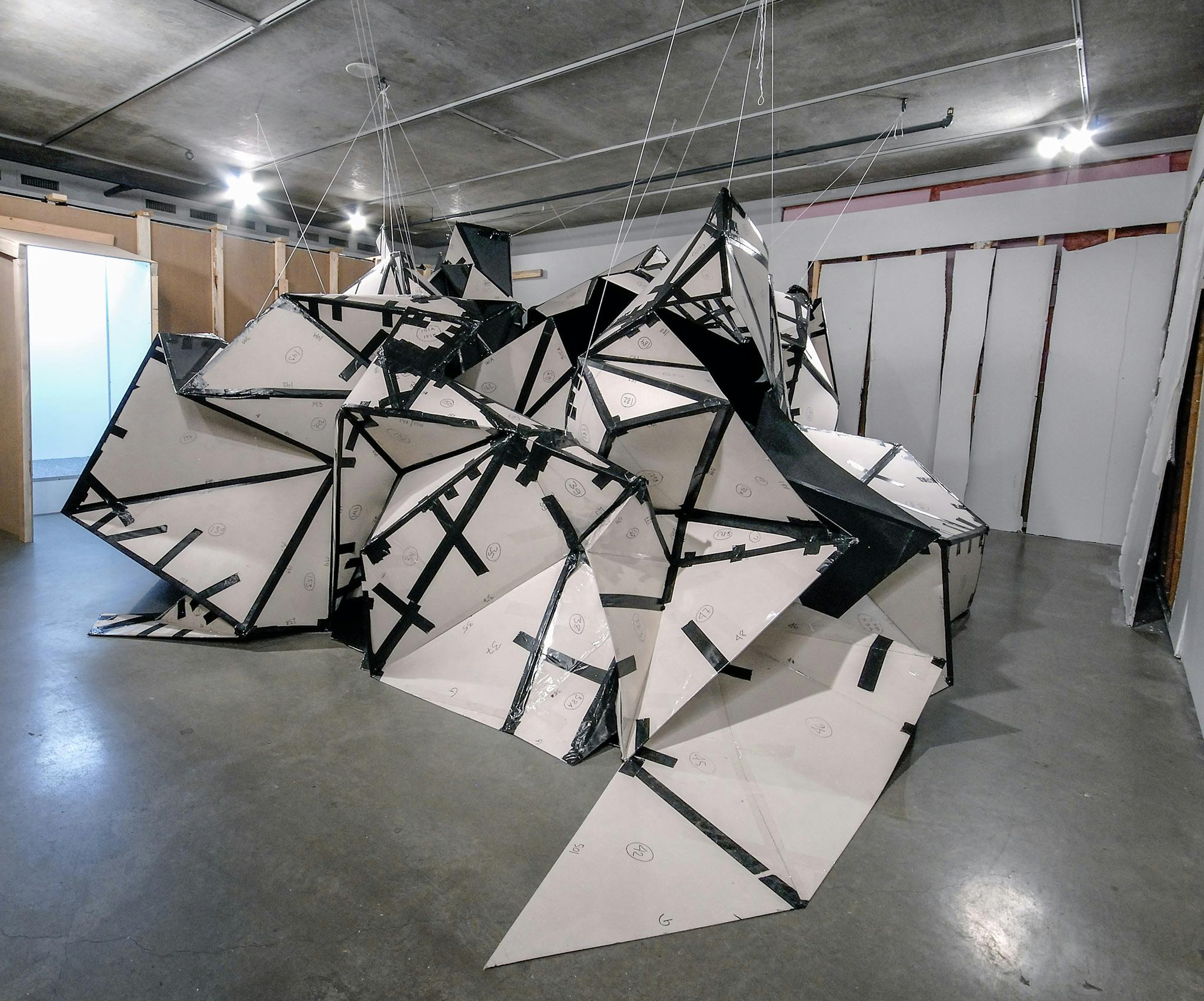 A large white and black sculpture is placed on a gallery floor. It is partially hanging from the ceiling via transparent wires. The sculpture resembles a half-completed paper origami. 