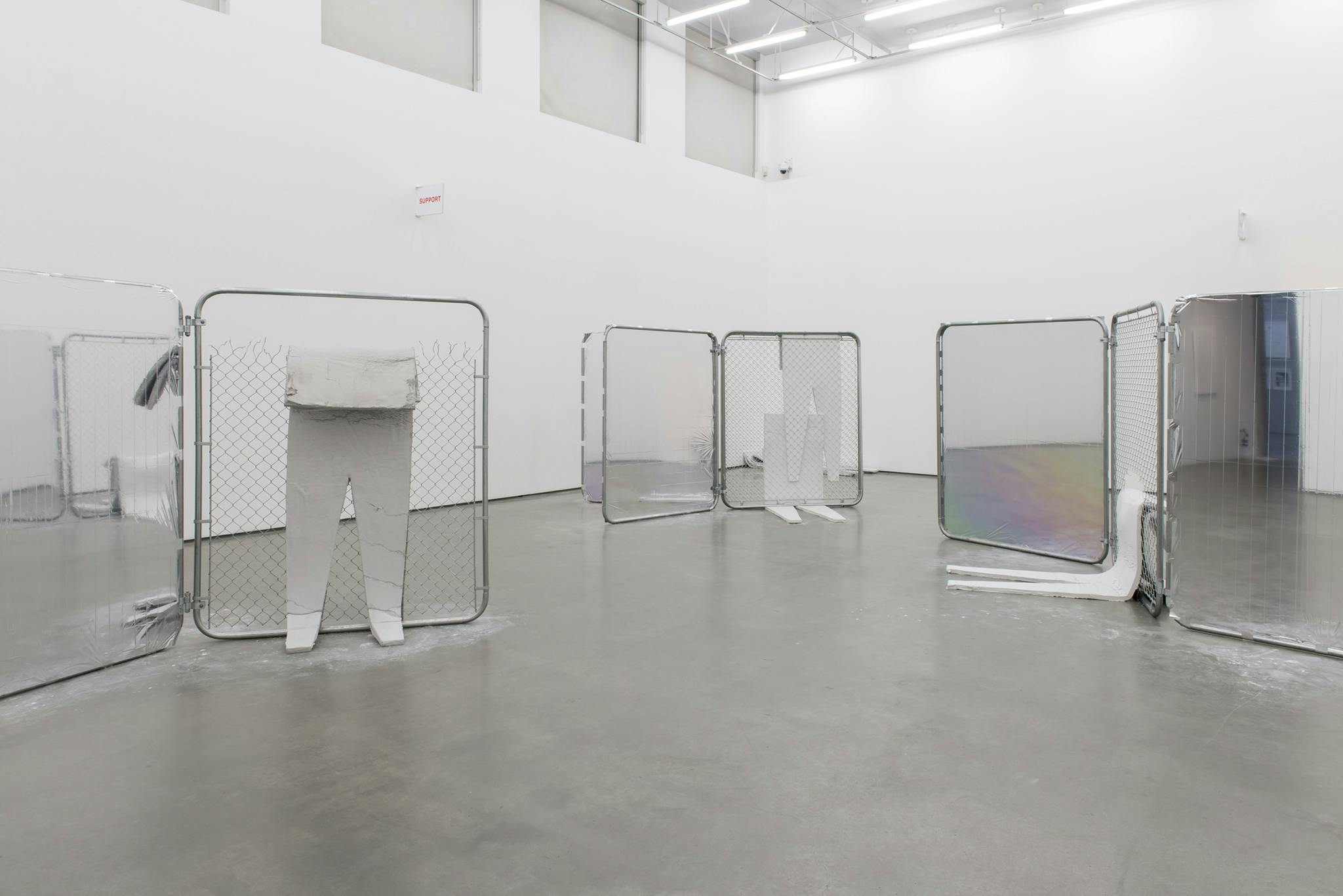 Various chain link fences sit staggered within a gallery space. Some have emergency blankets stretched taut in replace of chain links while thin, foam cutouts in human-like forms lean against others.