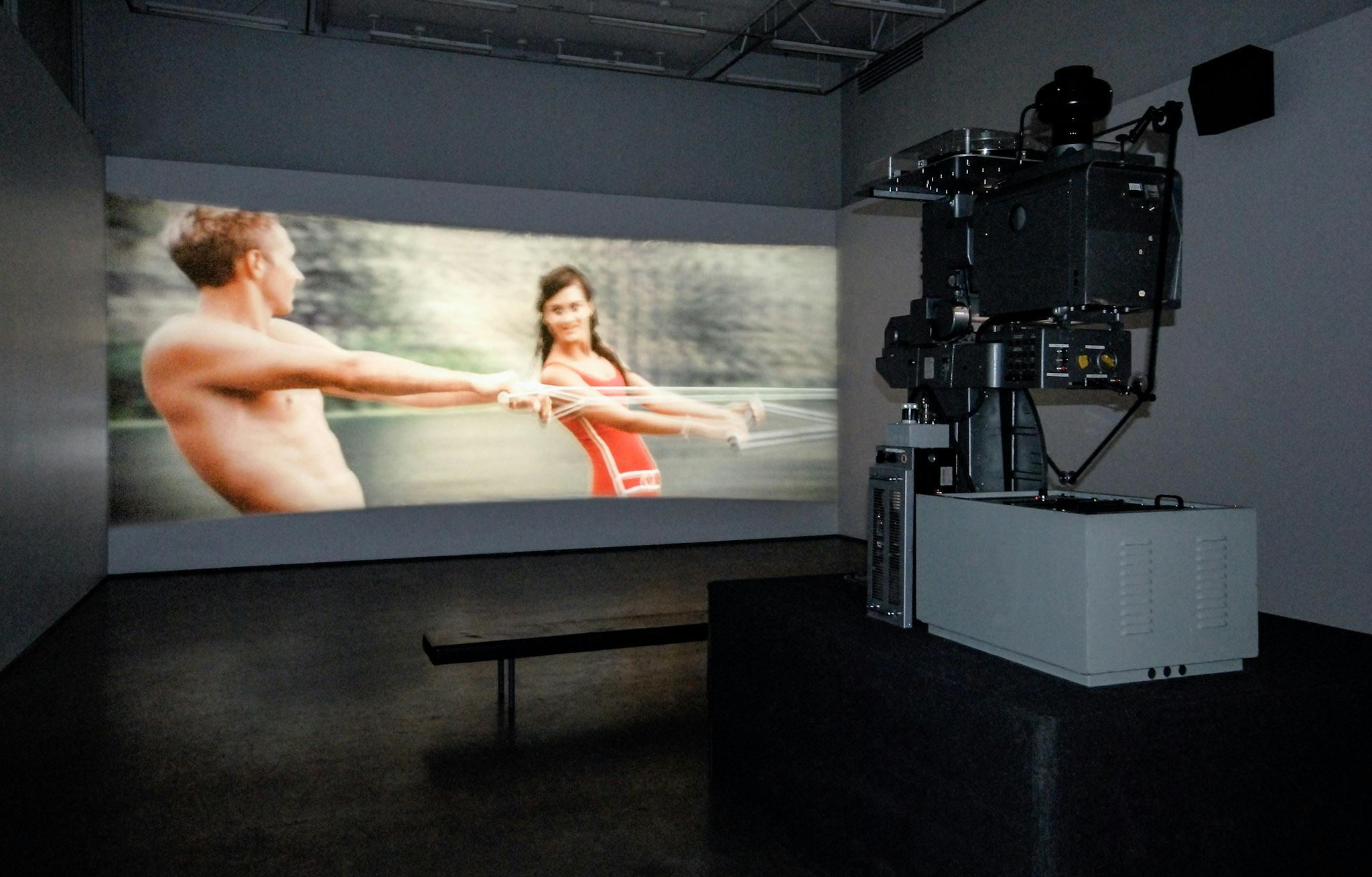 A film is projected on the wall in a gallery. The film depicts a man and woman in swimsuits. A silver film projector placed on the floor close to the gallery entrance projects this film to the wall. 