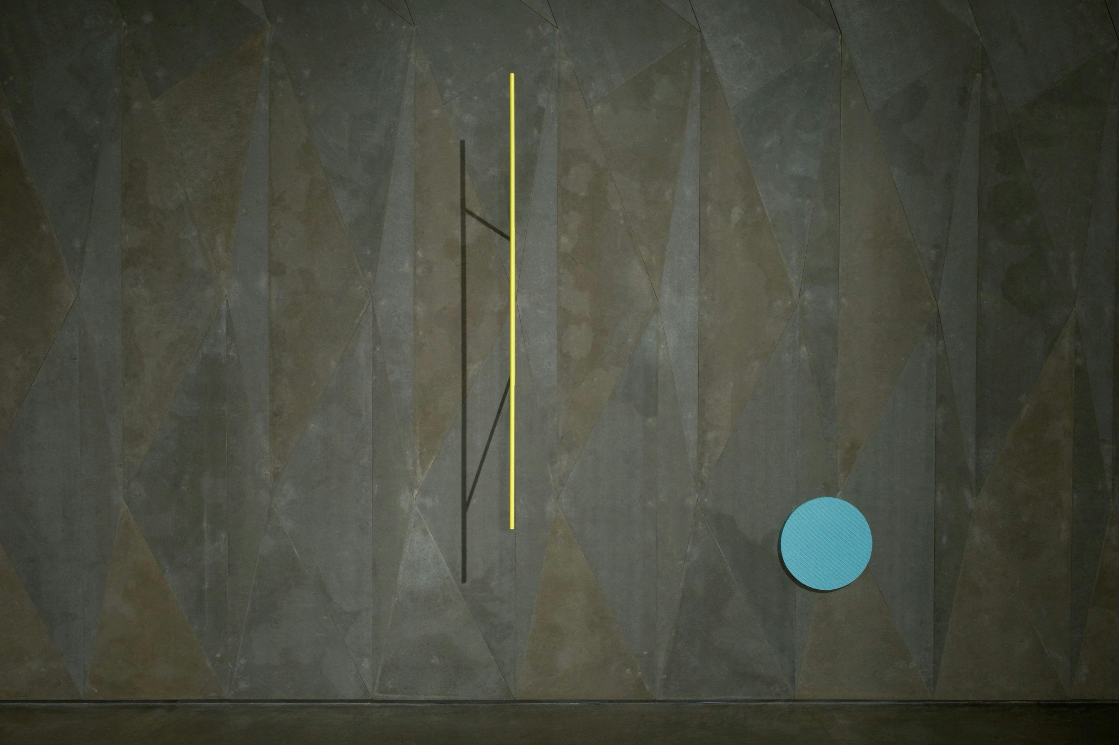 A textured, painted wall in gray on which two objects hang. One a sky-blue circle, the other a thin yellow stick hung vertically and casting a thin shadow to its right. 
