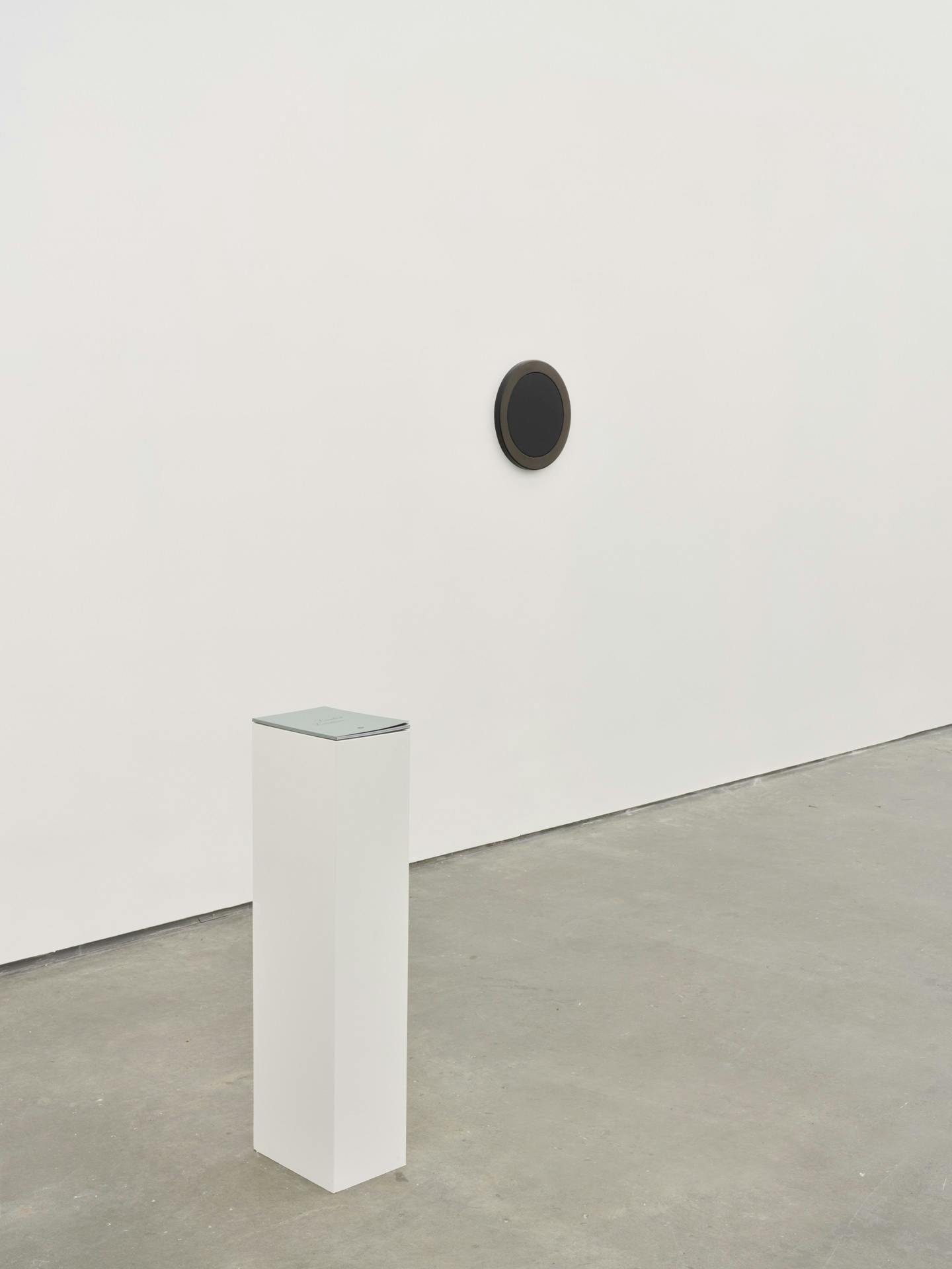 A thin artist book by Kathy Slade lying closed on a white plinth, in front of a white wall with a small, black framed mirror slightly to the right of the plinth.