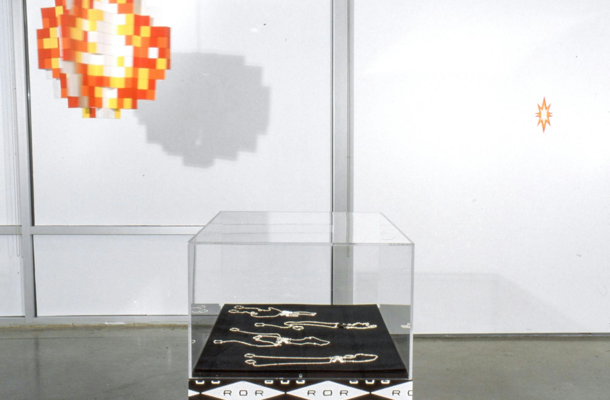 Installation image of works in a gallery. Four silver necklaces are placed flat on a piece of black fabric in a display case. A round sculpture that is probably made with orange, yellow and white Lego blocks is hanging from the ceiling.