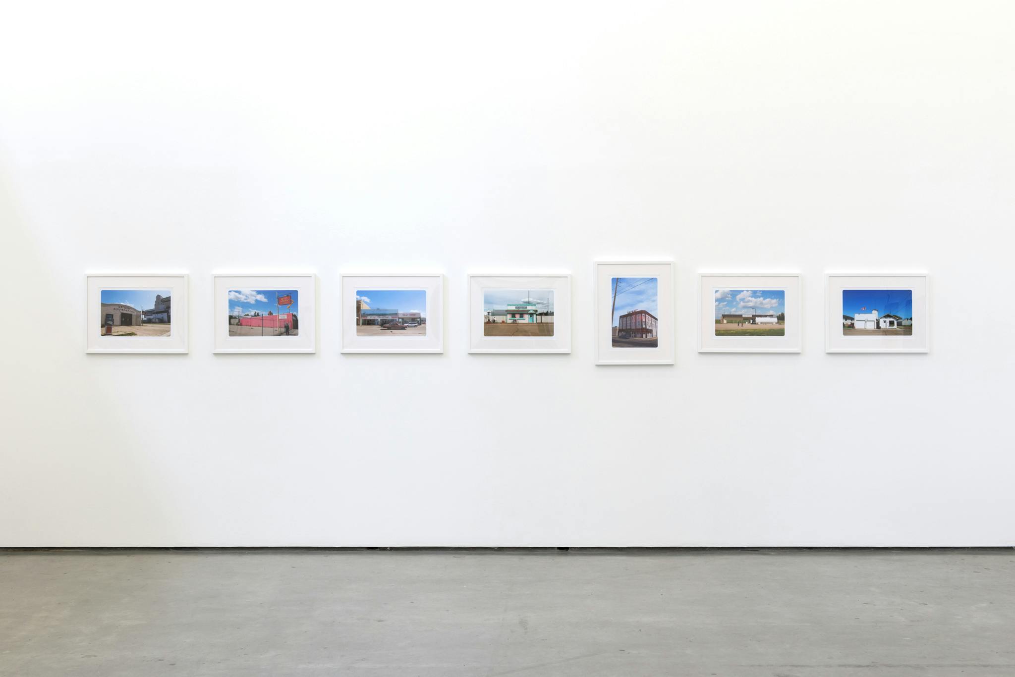 Seven framed photographs hang in a horizontal line on the wall of a gallery. The images depict various Saskatchewan rural buildings with signs in Cree syllabics, all with a blue sky above. 