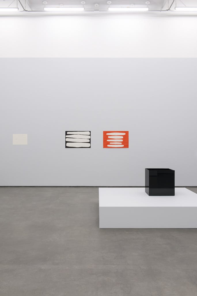 Jürgen Partenheimer’s artworks installed in a gallery space. A black, cube-like sculpture is installed on a low white plinth. Two abstract, watercolour paintings are installed on the wall behind.