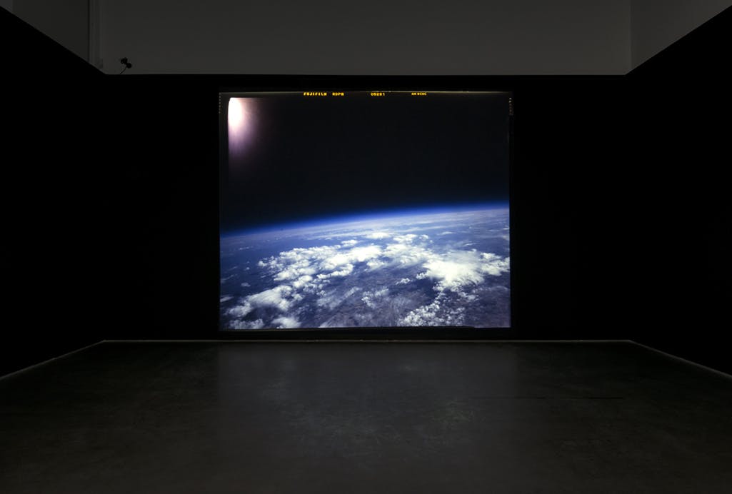 A picture of the earth is projected on a large screen on a black wall. The image depicts a view of the earth from space. Glare from the sun is visible at the upper left corner of the image. 