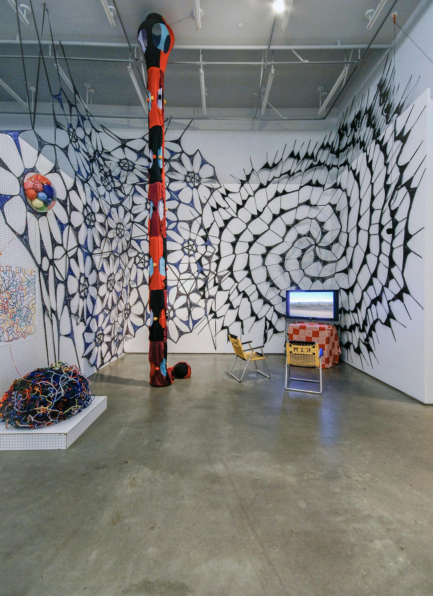 Various sculptures are installed in a gallery space. Large black web-shaped sculptures cover the gallery walls behind a TV monitor. A red tube-shaped sculpture is hanging from the ceiling.   