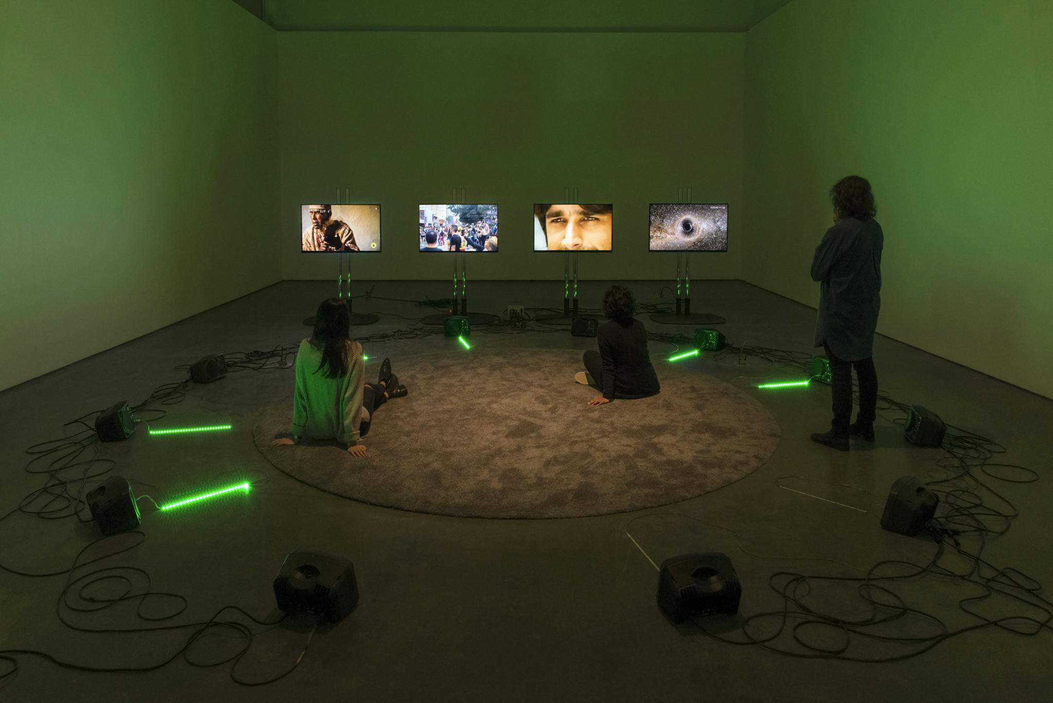 In a darkened gallery space, four thin TV monitors with stands play video works. In front of them, twelve speaker boxes are placed in a large circle, inside which visitors sit while watching videos.